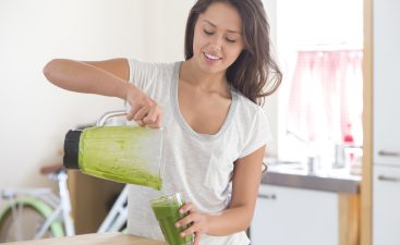 A happy young woman smiles as she pour a healthy homemade smoothie into a glass. She is standing in her kitchen wearing a plain white tshirt. A bike can be seen out of focus in the background.
